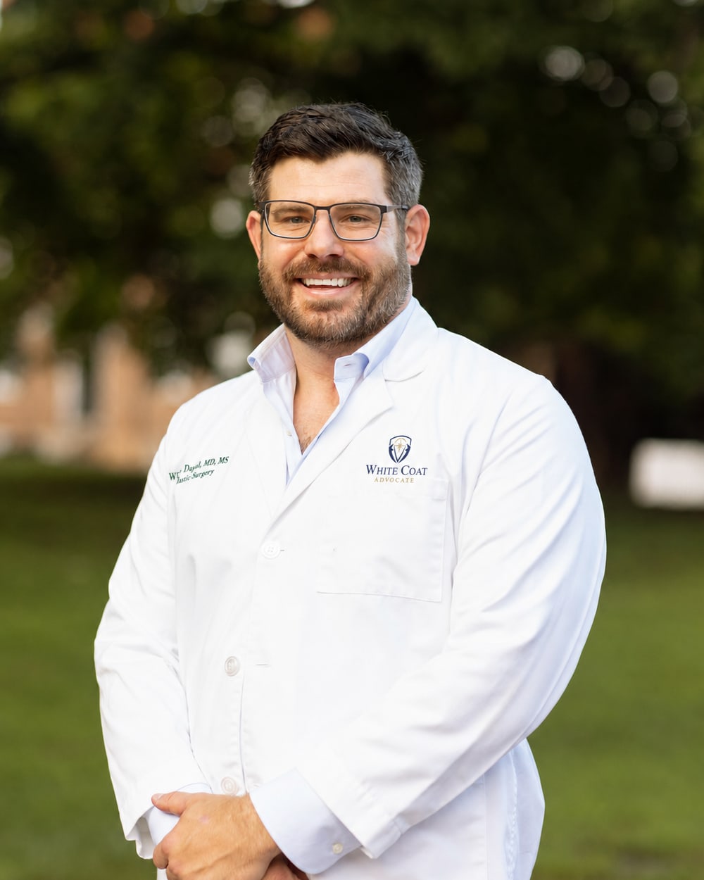 William Dugal, MD, standing outdoors in a white coat embroidered with the White Coat Advocate logo