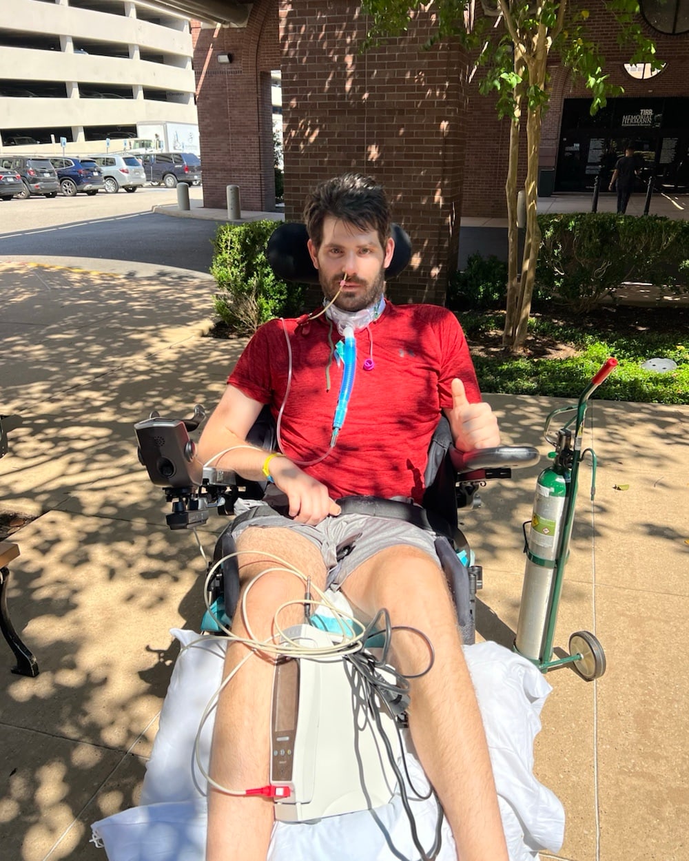 William Dugal outdoors in a powered wheelchair with oxygen and respiratory support during early recovery
